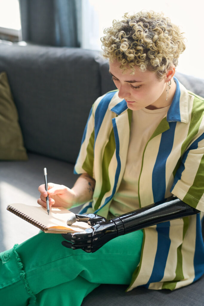 Young female with disability in casualwear sitting on comfortable couch in living room and drawing sketch with pencil in notepad